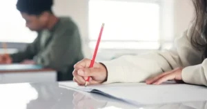 Primer plano de manos de estudiantes escribiendo con lápiz en cuadernos en un salón de clases de inglés.