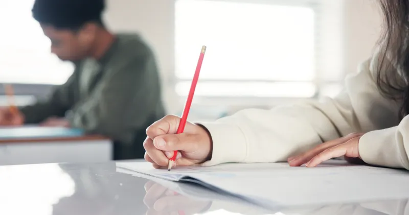Primer plano de manos de estudiantes escribiendo con lápiz en cuadernos en un salón de clases de inglés.