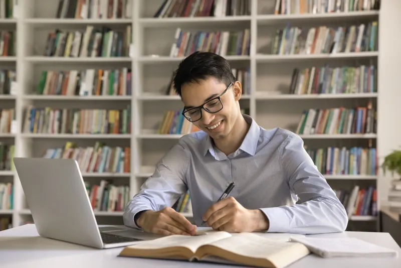 Joven profesional con gafas escribiendo en cuaderno mientras estudia inglés frente a un portátil en biblioteca.