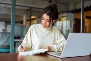 Mujer joven con suéter beige escribiendo en un cuaderno frente a su laptop en una oficina.