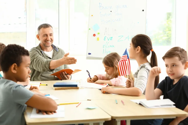 Profesor sonriente enseñando inglés a niños diversos en un aula con bandera de Estados Unidos.