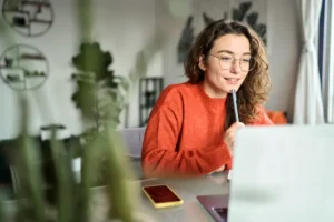 Mujer joven con gafas y suéter naranja estudiando inglés frente a un ordenador portátil en casa.