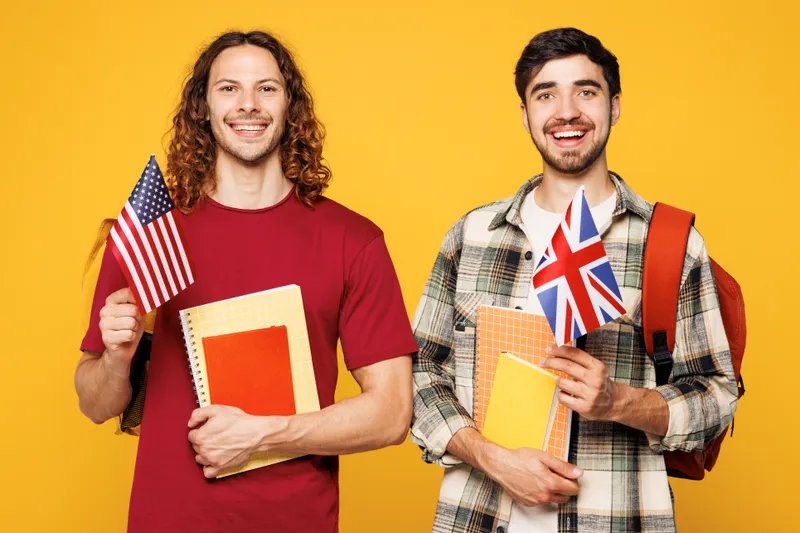 Dos estudiantes universitarios sonrientes sostienen banderas de EE. UU. y Reino Unido sobre fondo amarillo brillante.