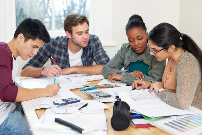Grupo multicultural de estudiantes jóvenes sentados alrededor de una mesa estudiando con libros y calculadoras.