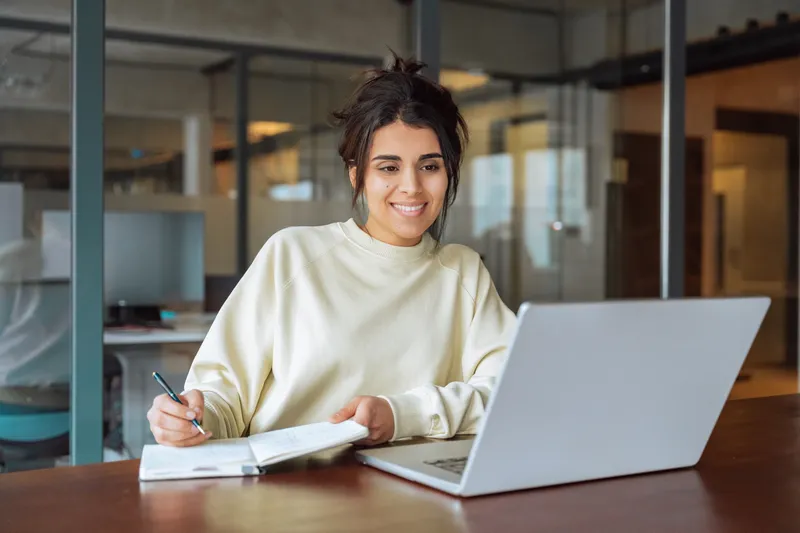 Mujer joven con suéter beige sonriendo mientras toma apuntes frente a su computadora portátil en una oficina moderna.